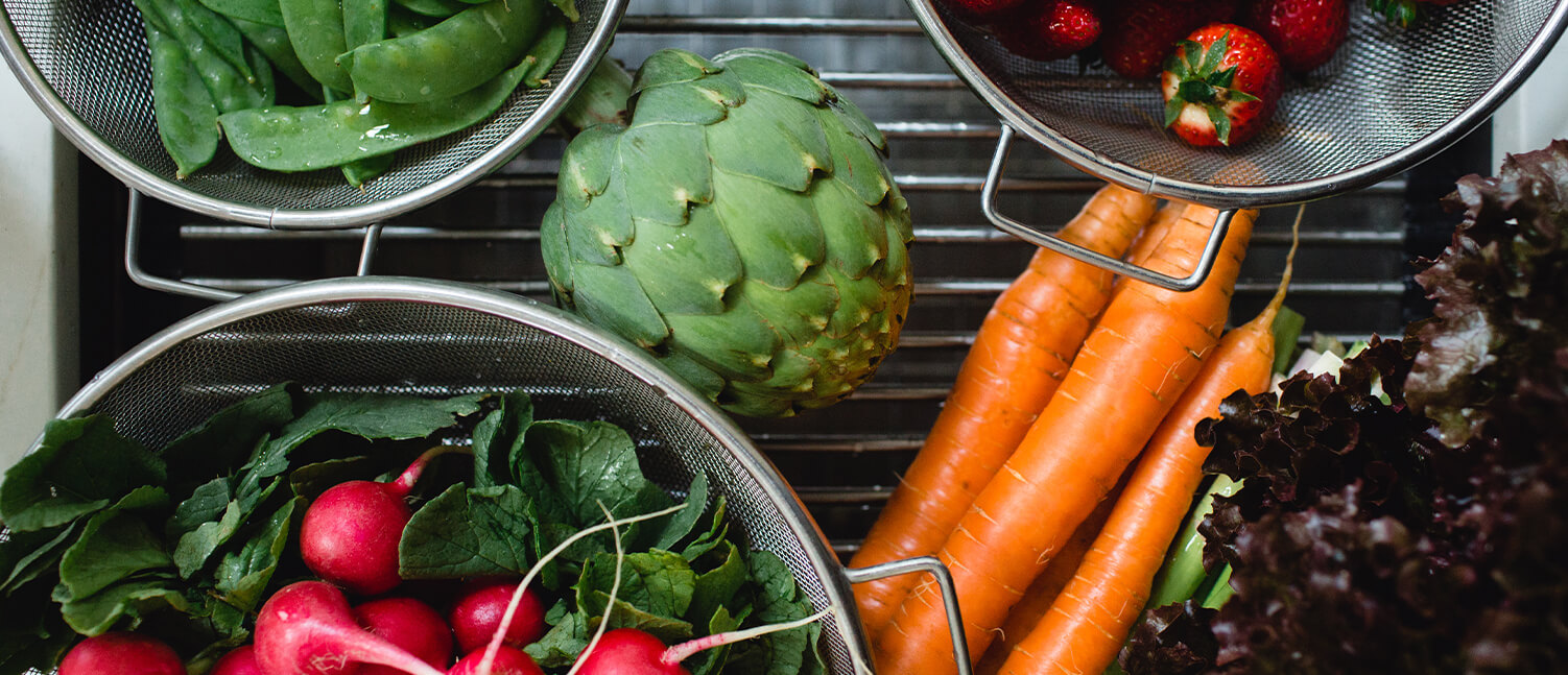 fresh vegetables being washed in a sink in colanders including radishes, lettuce, artichoke, carrots, peas, and lettuce.