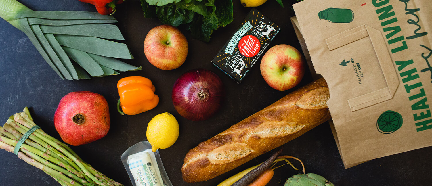 groceries falling out of a paper grocery bag including a baguette, fruits and vegetables.